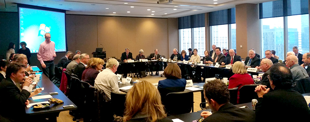 A large group of people seated at tables arranged in a square in a meeting room.