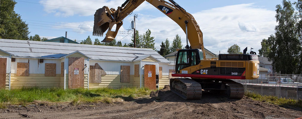 Photograph of a boarded-up home in the process of being demolished with heavy machinery.