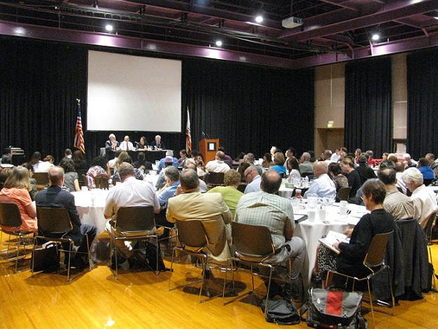 Photograph taken from the rear of a conference room of four presenters seated at a table on a stage in front of approximately 60 people seated around tables.