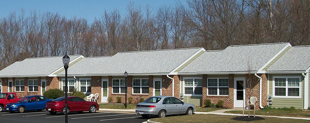Photograph taken at street level of the Cottages at Chesapeake showing five of the townhouses in the development. The one-story units have pitched roofs covered with shingles and facades of brick and wood siding. In front of the units is a parking lot. A sidewalk and narrow lawn area with shrubs and trees are between the building and the parking lot.