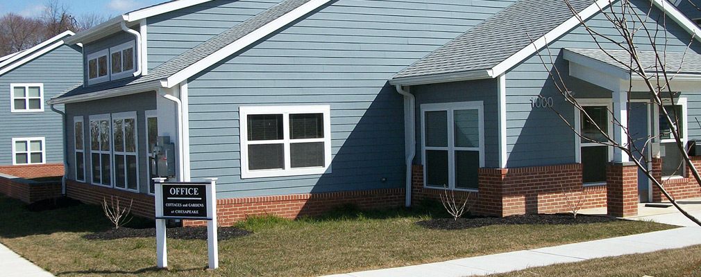Photograph taken at ground level of two sides of a one-story building. The entrance is on the gable end of the building. The facades are made of wood siding above a low brick base course. A shallow lawn with shrubs is on the front and side of the building. 