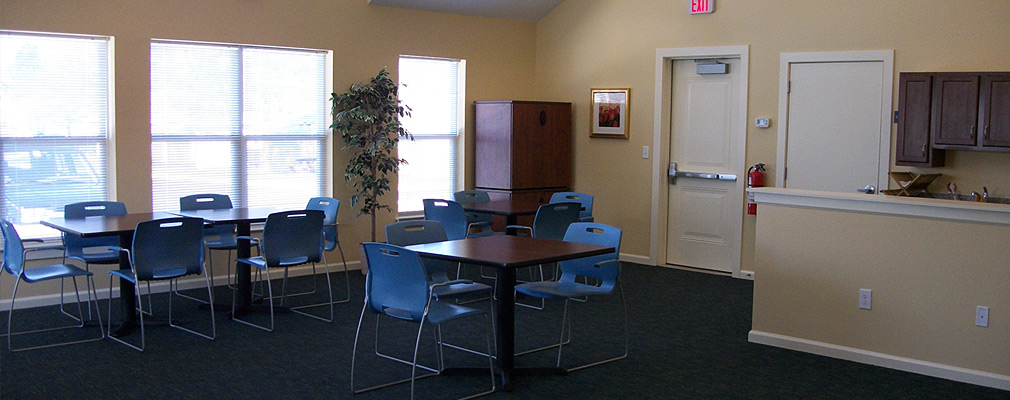 Interior photograph showing a corner of a meeting room in the new community center. Four small tables with chairs arranged in the room, which has a high ceiling and carpeting. A kitchenette is along one wall, and  large windows take up most of the other wall in the picture.
