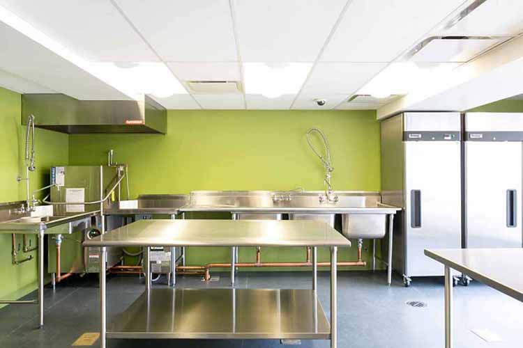 A photograph of the teaching kitchen showing preparation tables in the foreground, a sink along the left-side wall, and three sinks and two warming cabinets along the wall in the background. All fixtures are stainless steel.
