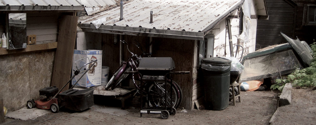 Photograph taken from street level of a one-story wooden house with a metal roof. A lawnmower, grill, trashcan, and two bicycles, are scattered near the entrance to the house. Elsewhere on the lot are a trashcan and an overturned wooden boat.