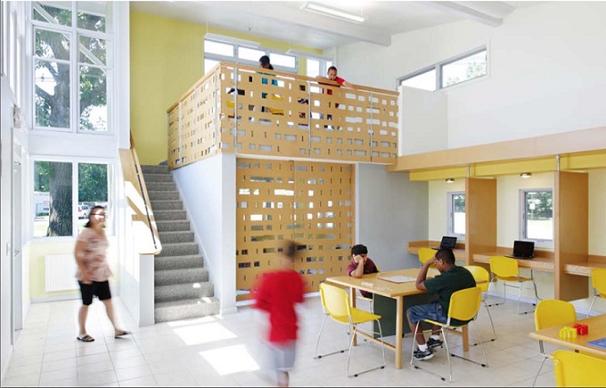 A photograph of the inside of the center’s large room. An adult is walking inside the building entrance. A child walks in the central portion of the room, where two other children are studying at one of tables. Three desk carrels line the side wall. Behind the children are a small room and stairs leading to the loft, occupied by four children.