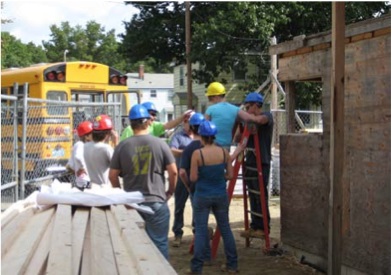 A photograph of nine instructors and students, all wearing hardhats, in the construction zone, with lumber in the foreground and a partially constructed wall near the students. A school bus is in the middleground behind a chain-link fence.