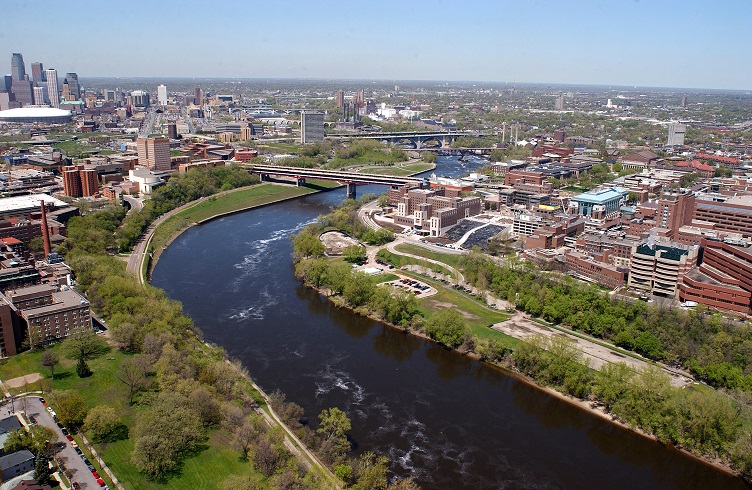 Low altitude aerial photograph, looking northwest, of the Minneapolis campus of the University of Minnesota straddling the Mississippi River. The skyline of downtown Minneapolis is in the middle ground at the left edge of the photograph.