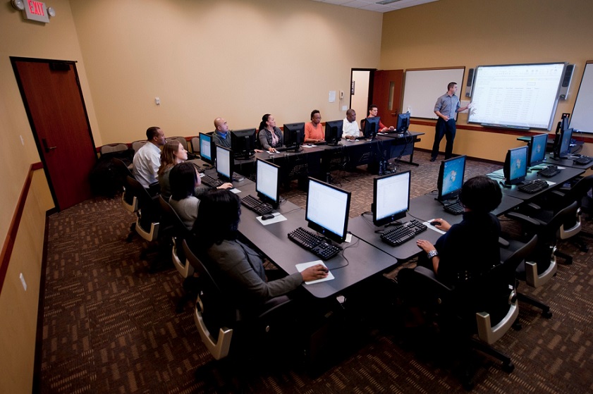 Photograph taken from an elevated position of a computer room, with 14 computers and 10 students at a U-shaped table. At the open end of the table, an instructor is pointing to an enlarged projection of the computer screen.