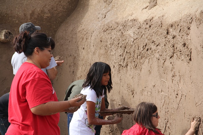 A photograph of six women, men, and children mud plastering an adobe home (courtesy of Tanya Hammiddi).