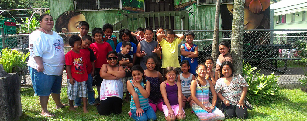 Photograph of an adult and elementary-school children in front of a one-story building. The front façade of the building is painted with a mural, with the profiles of two people’s faces set within tropical vegetation. 