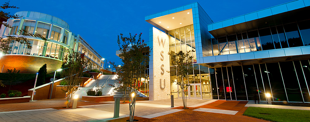 Photograph of the Winston-Salem State University’s student union building, shown in the evening, from the perspective of the pedestrian walkway in front of it. The walkway is planted with young trees. The two-story building’s façade is mostly glass; and near the entrance, a solid panel extends to the height of the building, with “WSSU” written vertically in large letters (courtesy of WSSU Photo: Garrett Garms).