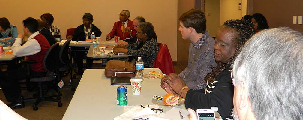 Photograph of approximately a dozen people seated at tables looking toward the front of the room. On the tables are sandwiches, sodas, and water bottles.