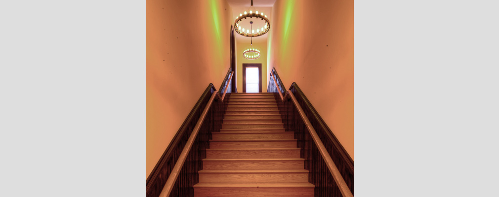 Photograph taken at the base of a long staircase, flanked by half-paneled walls and with a large two-tiered chandelier at the top of the staircase.  