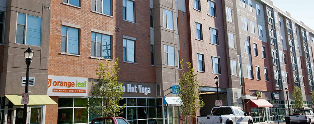 Photograph of the front façades of a block of five-story buildings with storefronts and a parking garage entrance on the first floor and residences on the upper floors.