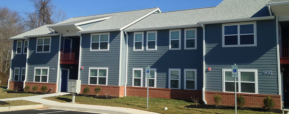 Photograph taken at ground level of the front façade of a two-story apartment building. The massing of the building is reduced by recessed sections of the façade, which also have slightly lower roofs. The facade is made of wood siding above a brick base course, and the pitched roof is covered with shingles.