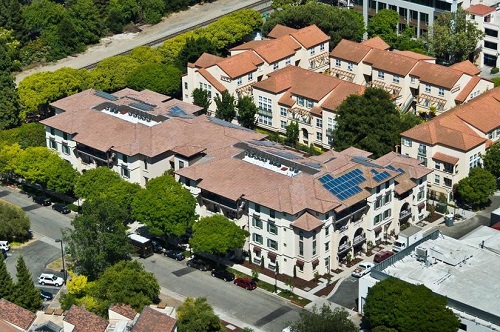 Low-angle aerial photograph of the four-story, c-shaped Franklin Street Family Apartments development. The building features a Mediterranean design and the roof includes solar photovoltaic panels. To the rear of the development are apartment buildings with a similar design.