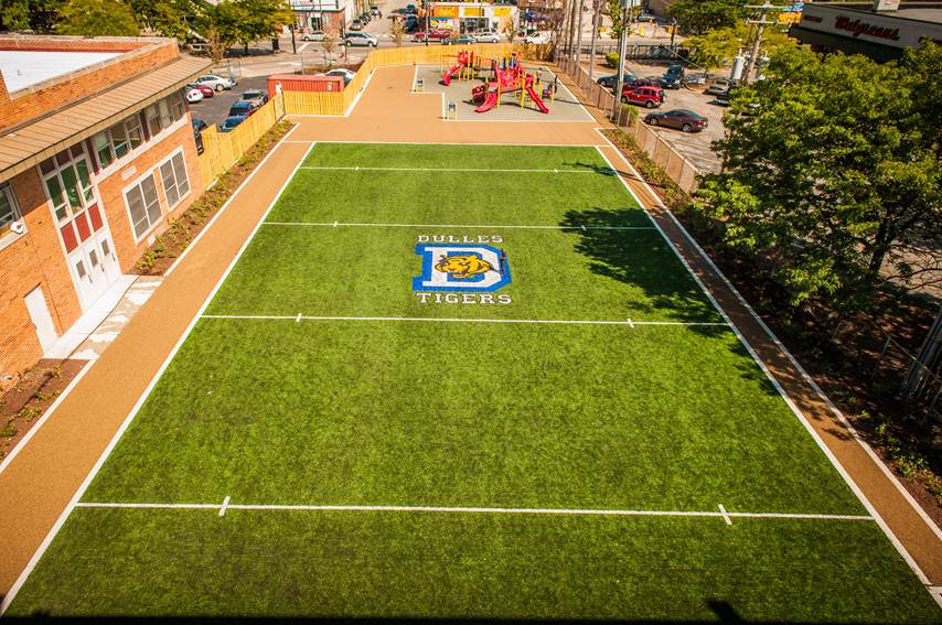 Low-angled aerial photograph of a mini football field. A school building and bushes are on one side of the field. Playground equipment is at the end of the field in the background.