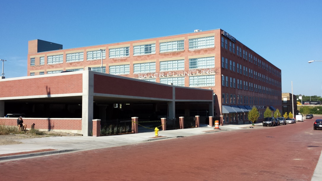 Photograph taken at street level from across the street showing two facades of a five-story brick building with a flat roof. “Baker Furniture” in faded paint is written above the third-story windows on one of the facades. In the foreground, a two-level brick-faced parking structure stands beside the converted industrial building.