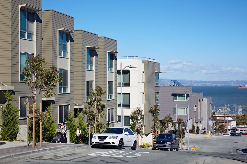 Photograph depicting multi-story residential buildings and the adjacent streetscape, including cars parked along the road and pedestrians traveling along the sidewalk. Views of the San Francisco Bay are visible in the background.