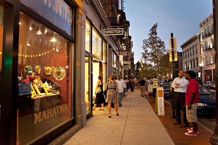 Photograph at dusk showing many pedestrians on the sidewalk in front of stores in historic buildings.