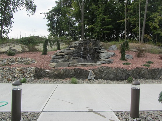 Photograph of water cascading down two levels of a rock water feature, which is surrounded with newly planted trees and groundcover. Large native trees stand at the top of the ridge behind the water feature. In the foreground is a sidewalk and two low light standards.