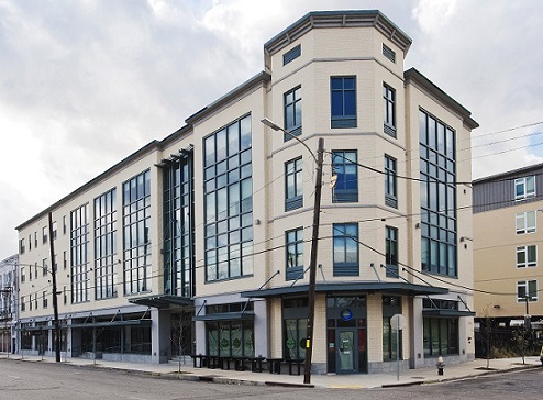 Photograph taken from across the street at ground level of the two street façades of the Harrell Building’s commercial wing. The four-story façade contains large windows on the first floor for the commercial spaces inside and many windows for the office space on the upper three floors. The building, standing immediately behind the sidewalk, has a flat roof and a three-sided corner articulation at the street intersection.