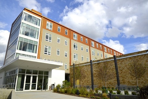 Photograph taken from ground level of two of the façades of King Rampart Apartments, a five-story building with a flat roof. The entrance at a corner of the building is marked by a cantilevered canopy and a glass door and walls. Small bushes line one side of the pathway to the entry.