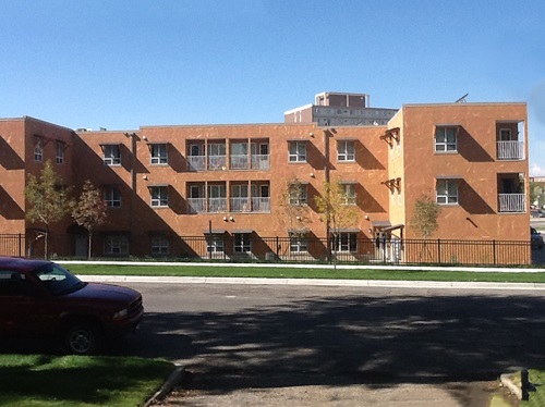 Photograph of a façade of a flat-roofed, three-story brick building, with a sidewalk, a street intersection, and a car in the foreground. An iron fence separates the property from the street right-of-way.