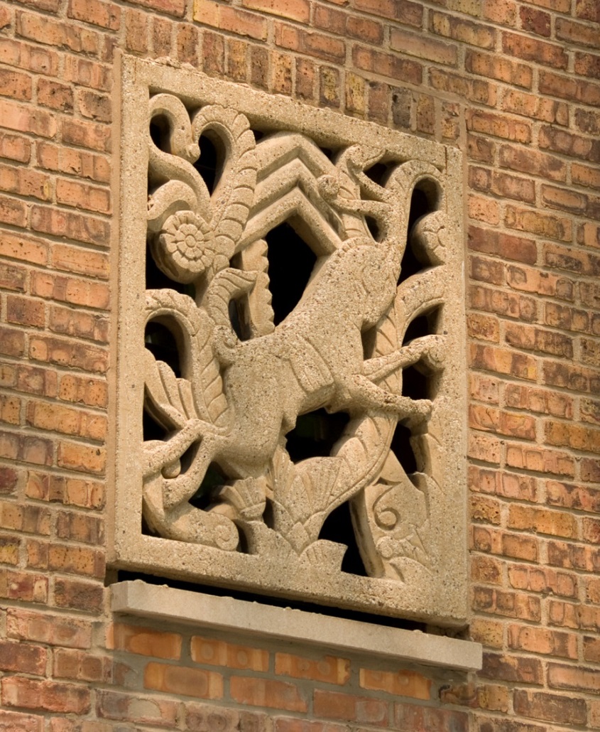 Photograph of a restored bas relief set in a wall of repointed brick. A running antelope is the central feature of the stylized sculpture, with geometric and natural shapes also set within the rectangular frame.