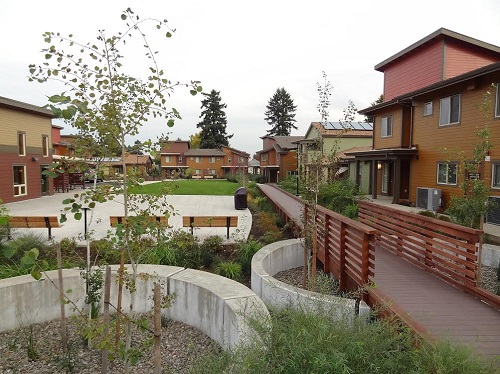 Photograph of several two-story buildings enclosing a landscaped courtyard that includes a lawn area, trees, a wooden walkway, and a paved area with several benches.