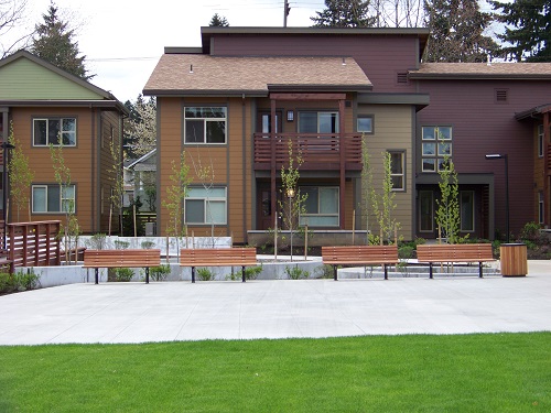Photograph of portions of the façades of two 2-story, wood-sided residential buildings facing the courtyard. A lawn and a paved open area with four benches are in the foreground.