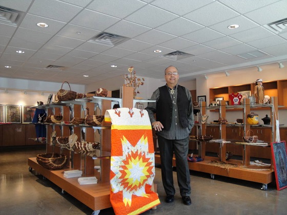 Photograph of the interior of the Turtle Mountain Tribal Arts Association retail space. Native American artisanal items are on display on two sets of shelves in the foreground; the store manager stands between the shelves. Paintings are visible in the background.