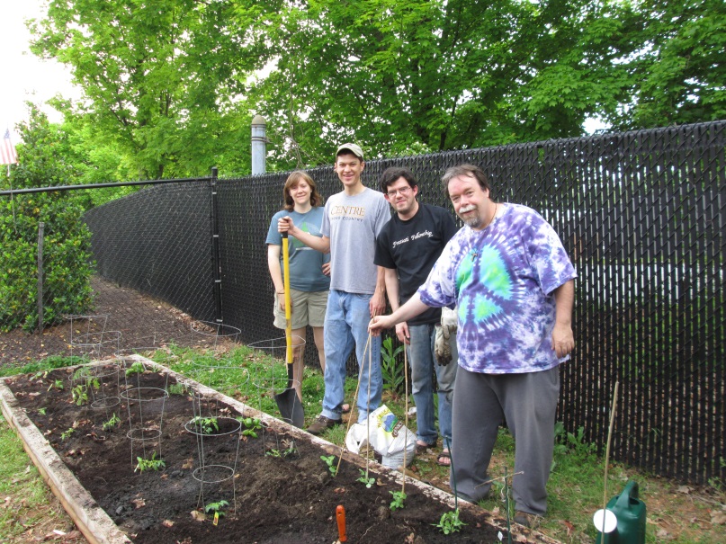 Photograph of three men and one woman standing beside a garden bed that they have been tending. About a dozen plants are sprouting in the bed that is edged by wooden four-by-four boards. Behind the gardeners is a slatted chain-link fence.