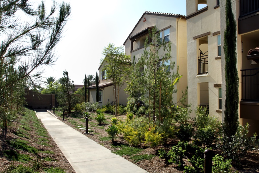 Photograph of a three-story building with stucco walls and a terracotta roof. Numerous shrubs and trees line a sidewalk in front of the building.