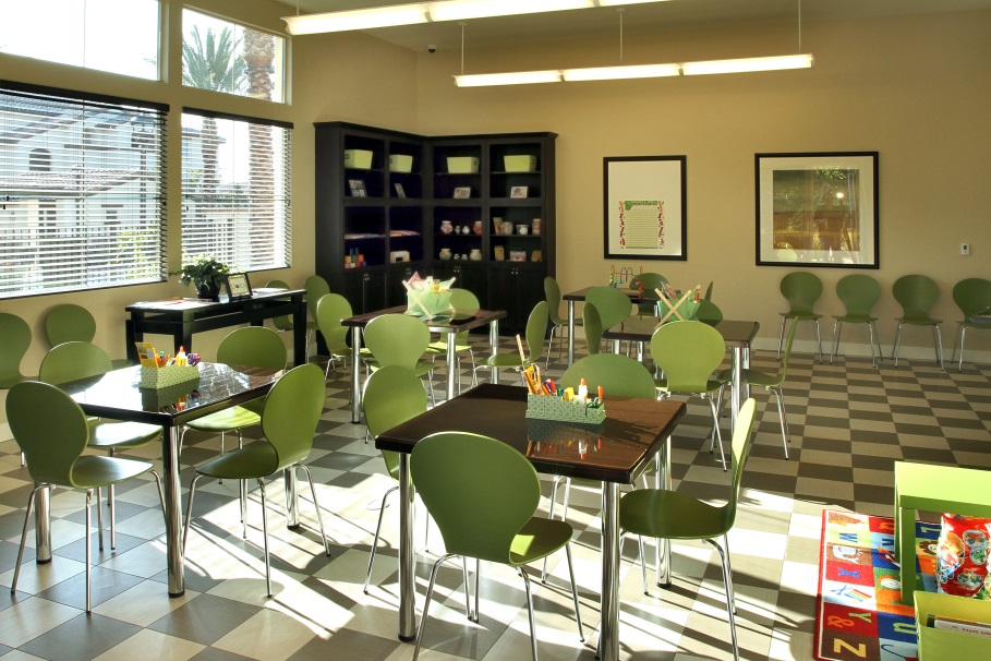 Photograph of a room with five work tables and four chairs at each table, a supply cabinet, and large windows.