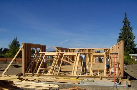 Photograph of four people participating in the construction of a home by lifting the framing of the fourth exterior wall into place.