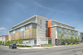 Photograph taken at street level from across the street at an intersection showing two facades of a four-story building with a flat roof. Portions of two similar buildings in the complex are in the picture on the far side of the two facades. The building is set back slightly from the sidewalk with a small area of bushes, grass, and street trees in the setback. 