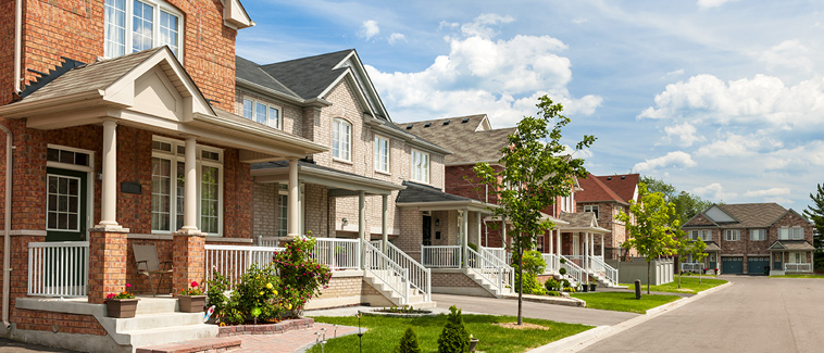 Photograph of several single-family homes, of a similar style but with varying facades, lining a street. Each of the homes has a front porch with stairs and a small yard with vegetation.