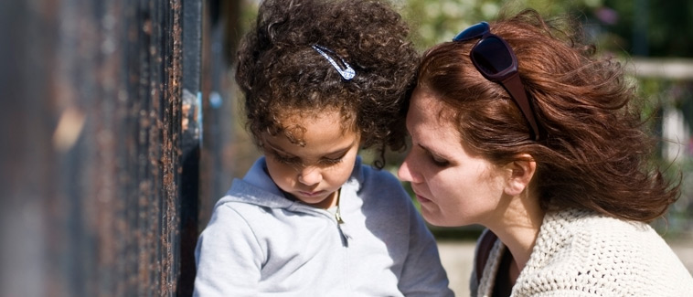 Image of a woman at eye-level with a young girl outside. Vegetation is visible in the background.