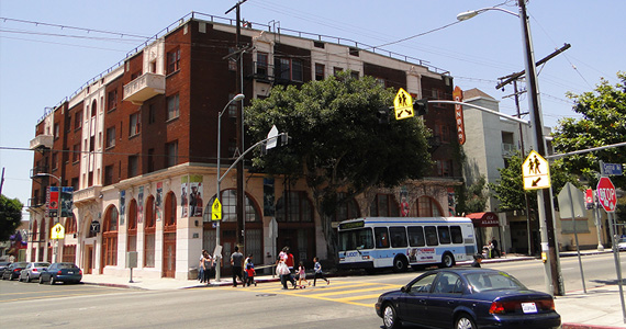 Photograph taken at street level of the Dunbar Hotel in Los Angeles. The building has a brick façade, large entry doors, and multiple windows. Trees in the foreground add aesthetic value to the building.
