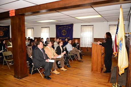 Interior image of the Peter DeBaun House, with several seated individuals listening to Rachelle Levitt, Director of the Research Utilization Division of the Office of Policy Development and Research at HUD, standing behind a podium speaking to finalists. The American flag, as well as American Legion flags, are visible in the photo.