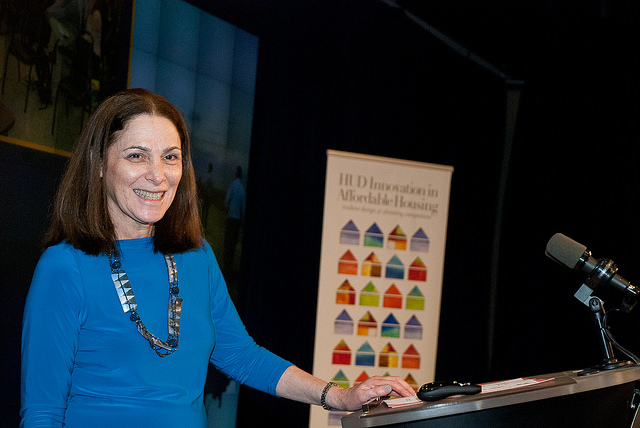 Photograph of Rachelle Levitt standing at a podium onstage. A poster for the Innovation in Affordable Housing Student Design Competition is visible in the background. 