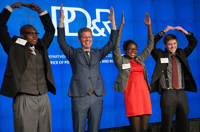 Four individuals, including three members of the Ohio State University team and HUD Secretary Shaun Donovan (second from left), pose in front of a backdrop with a PD&R logo, using their arms to spell out the word “Ohio.”