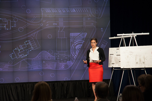 A student presenter stands in front of a screen displaying a site plan, next to posters mounted on easels. A seated crowd is visible in the forefront.