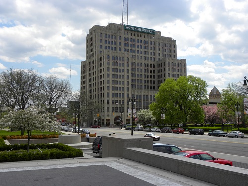 Image of a multi-story Wayne State University building, surrounded by vegetated landscape and fronting a multi-lane road with several parked cars.