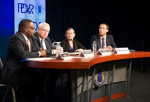Four individuals sit at a table on stage with name cards and microphones; the PD&R logo is visible on a screen in the background. 