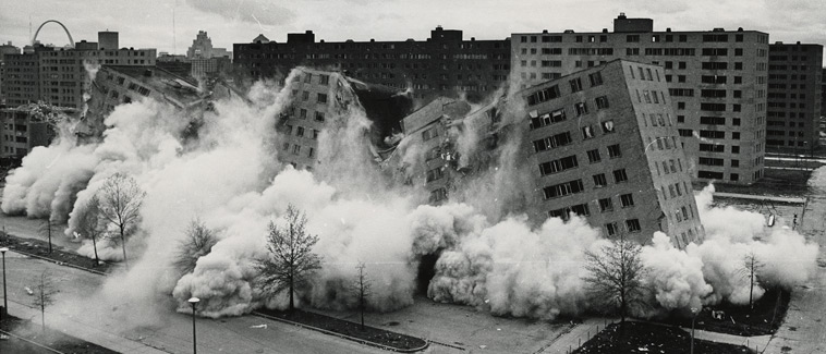 Image showing several towers of the Pruitt-Igoe public housing development in St. Louis being demolished. The high-rise, multi-story residential buildings are surrounded by parking lots, streets, and small strips of vegetation.