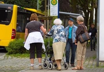 A senior woman with a walker and three other individuals gather at a bus stop, near a transit bus. 