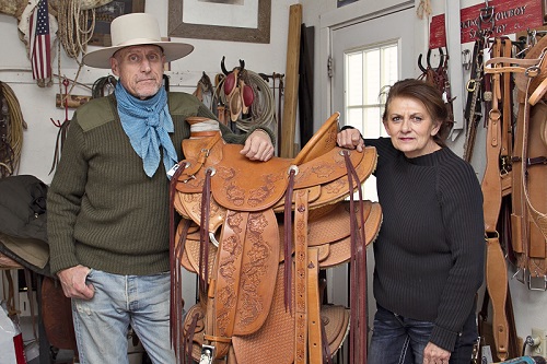 Photograph showing two individuals, a man and a woman, standing on either side of a stack of saddles on display.