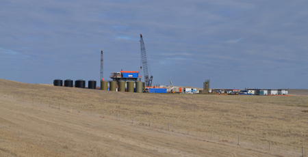 Photograph of oil recovery equipment, including a drilling rig and oil drums, in the middle of an empty field at the Bakken formation in Williston, North Dakota.